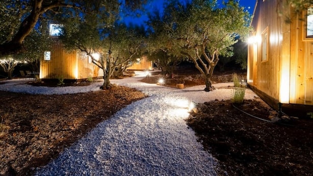 A pathway leading to wooden cabins with outdoor lighting, surrounded by trees and gravel.
