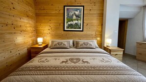 A bedroom with a wooden wall, a bed with patterned bedding, two bedside tables with lamps, and a framed picture above the bed.