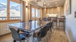 Una cocina moderna con armarios de madera, una amplia mesa de comedor con sillas, luces colgantes y vistas a montañas nevadas a través de grandes ventanales.