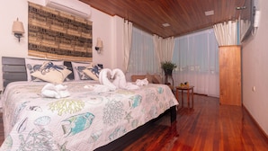 Une chambre avec un plafond en bois, un lit recouvert d’une couvre-lit à motifs, un climatiseur fixé au mur, une lampe de chevet et une petite table sur laquelle se trouvent des fruits.