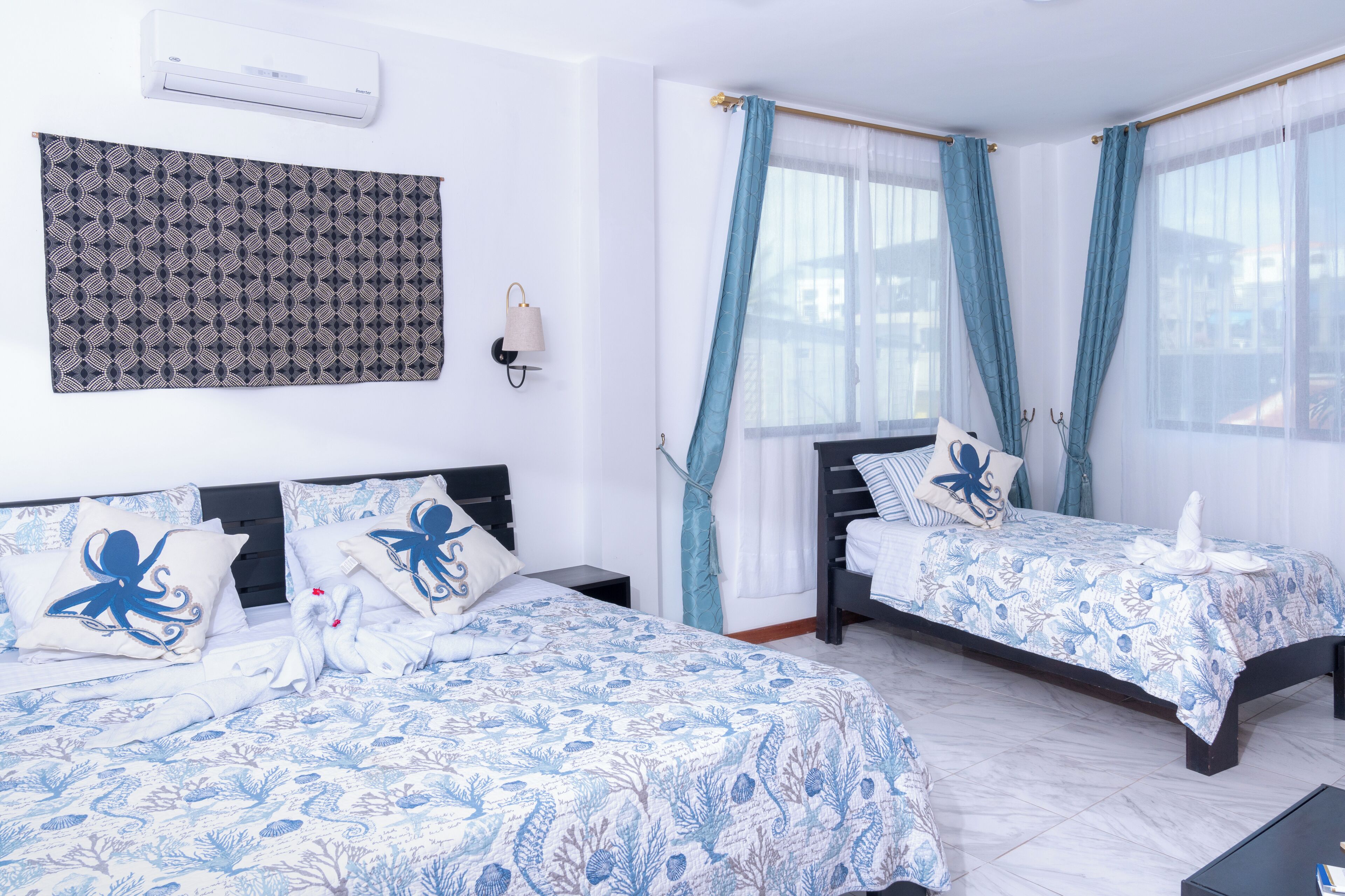 A hotel room with two beds, a patterned wall hanging, and a window with blue curtains.