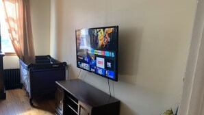 Un salon avec un téléviseur à écran plat fixé au mur, qui diffuse différentes chaînes. On aperçoit un meuble en bois foncé sous le téléviseur, et un radiateur est visible au fond de la pièce.