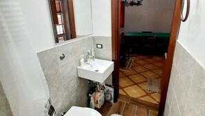Een kleine badkamer met toilet, wastafel en spiegel. Een deur leidt naar een eetruimte.