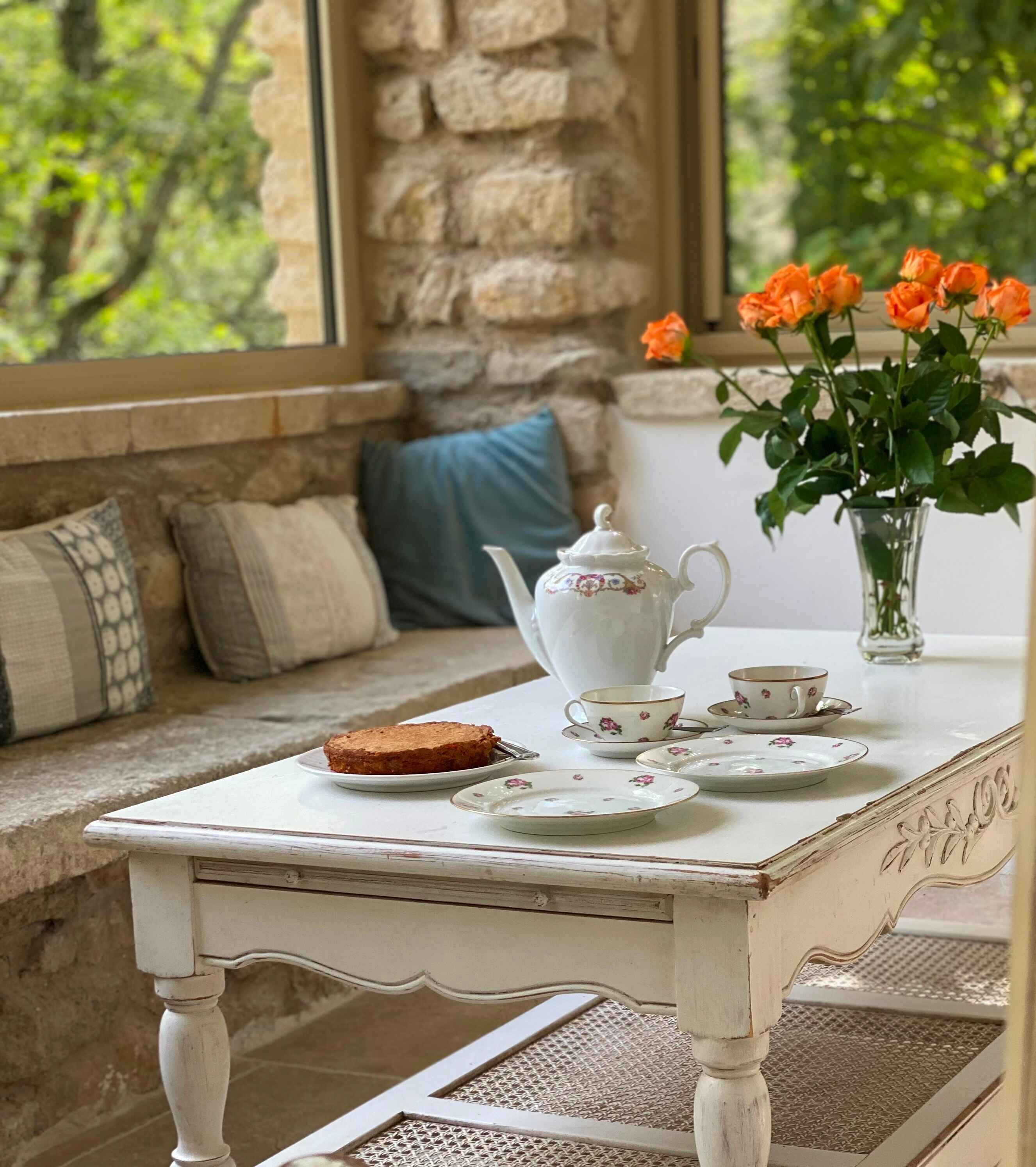A table set with a teapot, cups, saucers, and a plate with toast, placed on a window seat with a view of greenery.