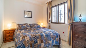 Un dormitorio con una cama, dos mesitas de noche con lámparas, un aparador, un ventanal con cortinas y un cuadro colgado en la pared.