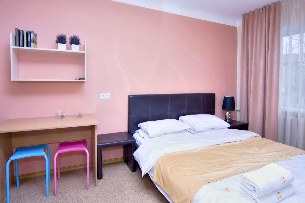 Quarto de hotel com cama, mesa de cabeceira, luminária, escrivaninha com duas cadeiras e estante com livros e plantas.