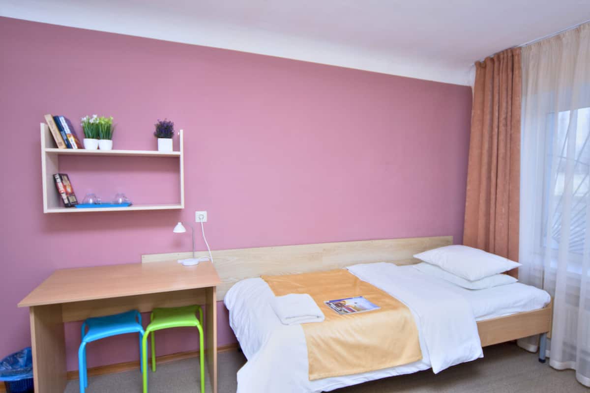 Um quarto com cama, escrivaninha, duas cadeiras, uma estante com livros e plantas, e uma janela com cortinas.