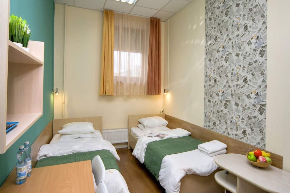 Quarto de hotel com duas camas de solteiro, uma mesa pequena com uma cesta de frutas, uma prateleira com garrafas de água e uma janela com cortinas finas.