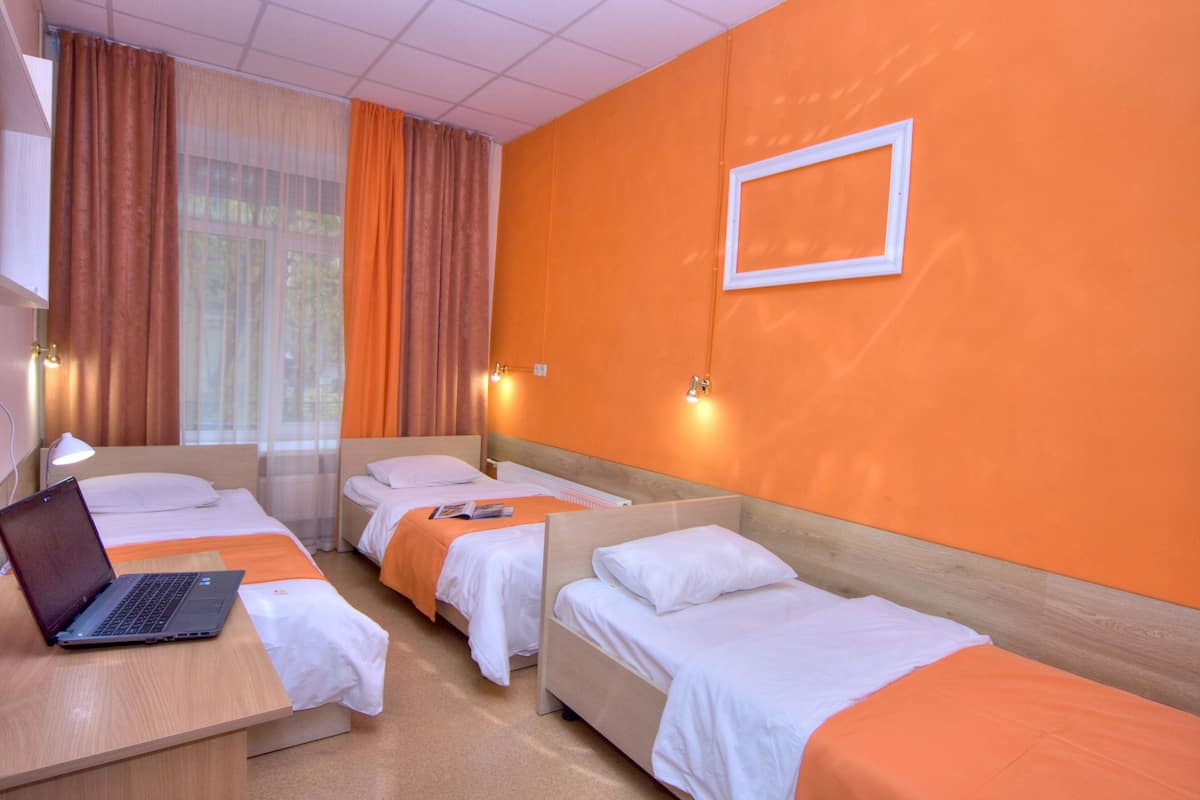 Quarto de hotel com três camas de solteiro, uma escrivaninha com um laptop e uma janela grande com cortinas.