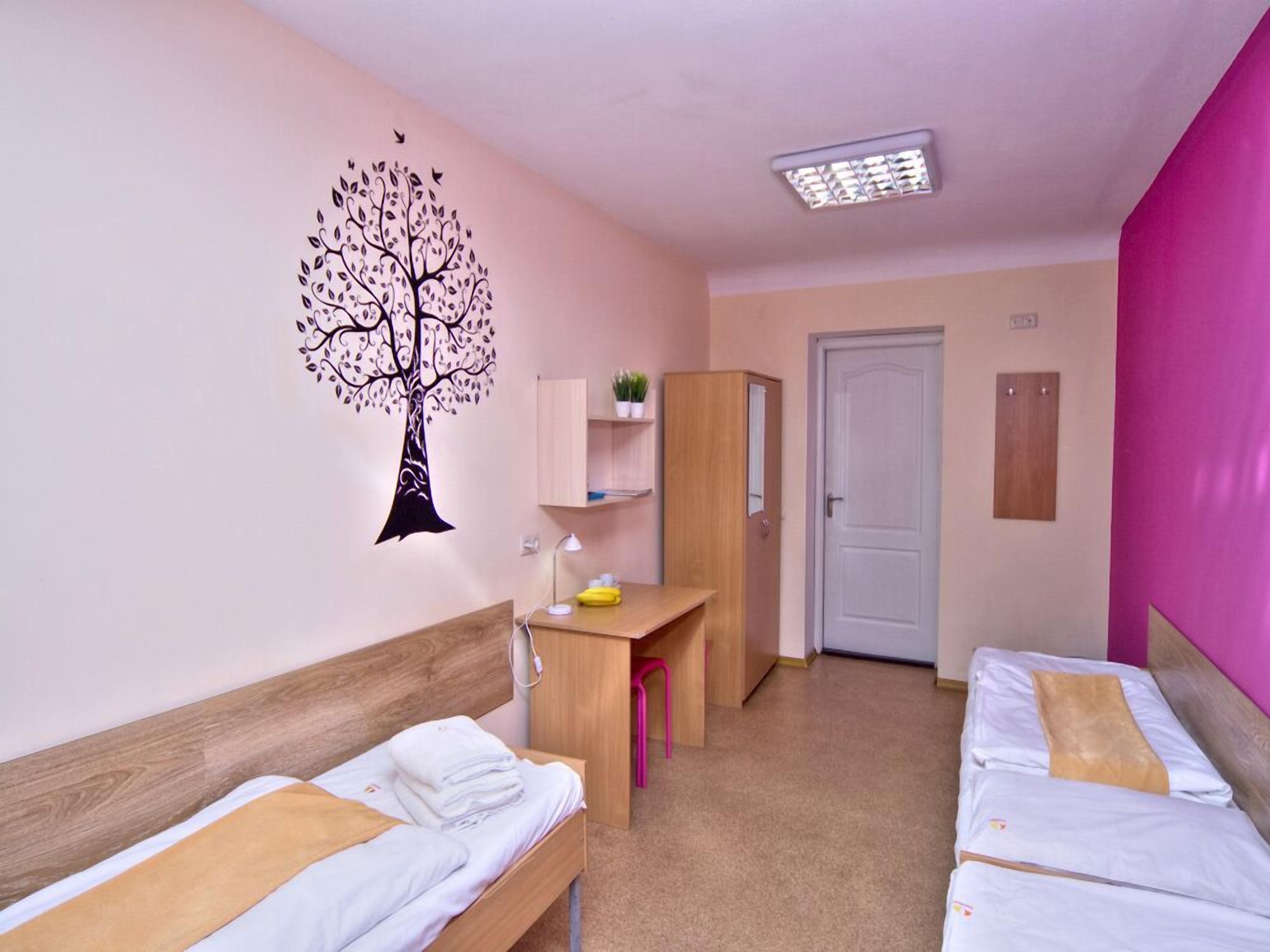 A hotel room with two beds, a desk, a chair, a wardrobe, a door, and a wall decoration of a tree.