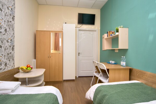 A hotel room with two beds, a small table with a fruit bowl, a chair, a desk with a shelf, and a TV mounted on the wall.