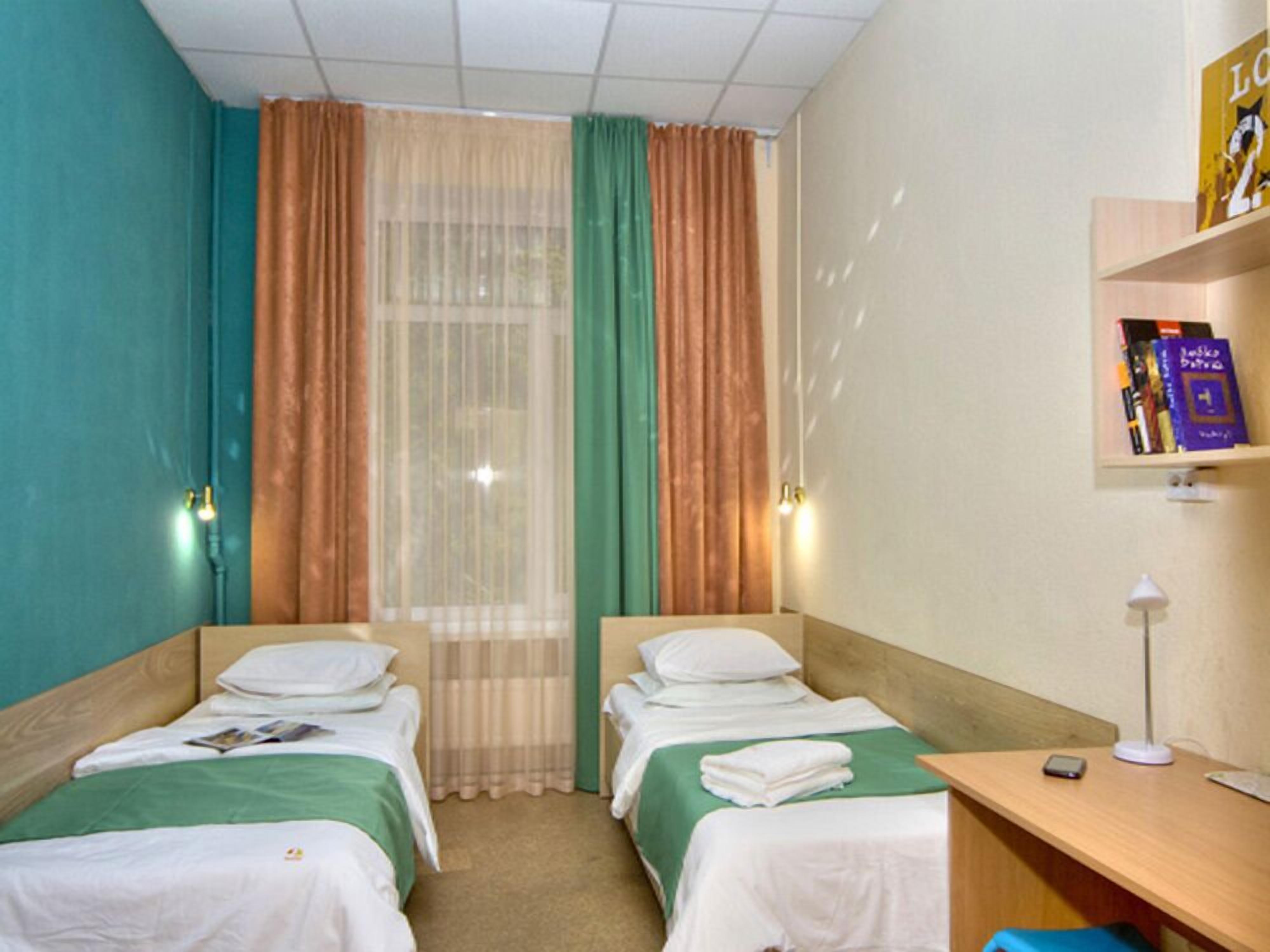 A hotel room with two single beds, a desk, a chair, a shelf with books, and a window with curtains.