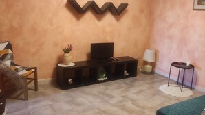 Een woonkamer met een televisie op een zwarte standaard, een vaas met bloemen, een lamp en een klein tafeltje met een rode beker.