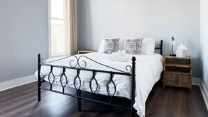 Camera da letto con un letto a struttura in metallo, biancheria da letto bianca, cuscini decorati, un comodino con una lampada e un tavolino di vimini.
