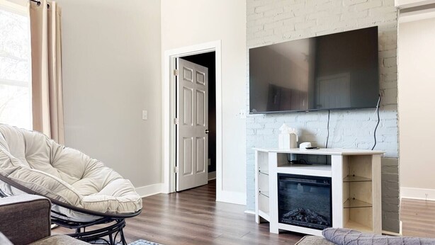 Un soggiorno moderno con parete in mattoni bianchi, una TV a schermo piatto montata sopra un mobile porta TV in legno, una poltrona grande e confortevole e un divano.