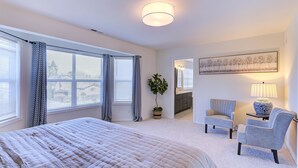 Une chambre avec un grand lit, deux chaises, une petite table, une plante en pot et un tableau accroché au mur.