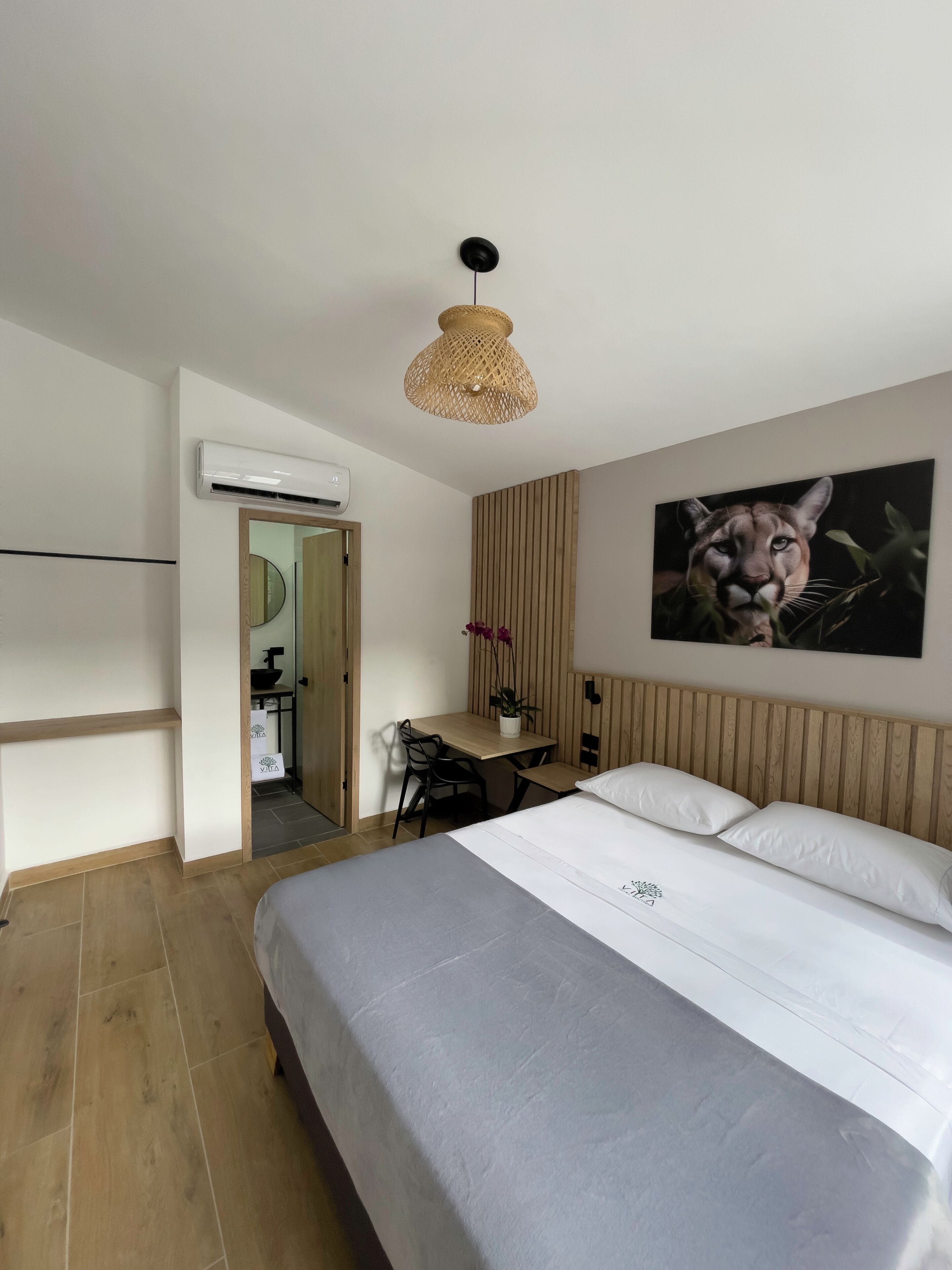 Ein Hotelzimmer mit Bett, Schreibtisch, Stuhl, einer Pendelleuchte, einer Klimaanlage und einem Wandbild.