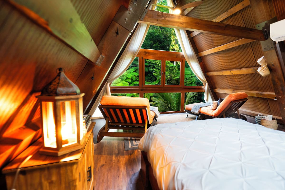 Une chambre confortable située dans les combles, avec une grande fenêtre, des poutres en bois, un lit, une chaise et une lanterne.