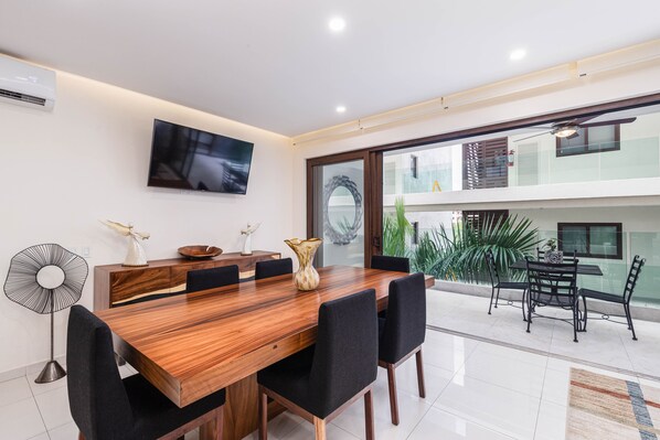 A modern dining area with a wooden table, black chairs, a TV mounted on the wall, and a large window offering a view of the outdoors.