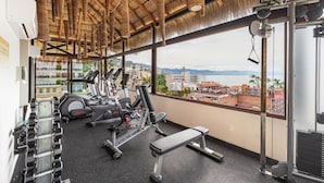 Un gimnasio con equipo de ejercicio, una banca y vistas a un paisaje urbano a través de amplios ventanales.