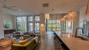 A modern kitchen with a long island, bar stools, and large windows offering views of the outdoors. - The Birch Bower: Luxury Lakefront Home (Oakland)