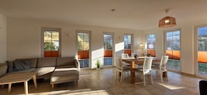 A spacious living area with a sofa, a dining table with chairs, and large windows with blinds.