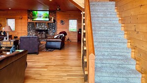 A spacious wooden interior with a staircase leading to an upper level, a stone fireplace, and a television in the living area. - Dramatic Remodeled Log home with everything! (Franklin)