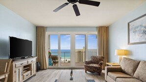 A hotel room with a balcony, a flat-screen TV, a wooden desk, a patterned armchair, a sofa, and a lamp.