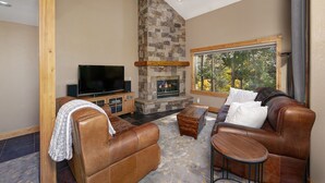 A cozy living room with a stone fireplace, leather sofas, a flat-screen TV, and a large window offering a view of trees. - Mountain Retreat Near Hiking and Golf In Silverthorne (Silverthorne)