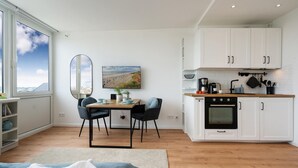 A modern kitchen with white cabinets, a wooden countertop, and a built-in oven. There is a dining area with a table and chairs, and a large window providing a view of the sky and buildings. - Himmelblau - Modern Renovierte, Zentrale Whg. Inkl. Pkw-stellplatz, Wlan. Haustiere Erlaubt (Westerland)