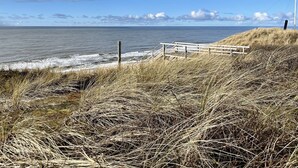 A coastal landscape with a wooden walkway leading to the beach, grassy dunes, and a clear view of the ocean. - Himmelblau - Modern Renovierte, Zentrale Whg. Inkl. Pkw-stellplatz, Wlan. Haustiere Erlaubt (Westerland)