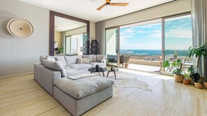 Un salón amplio con un sofá seccional gris grande, un ventilador de techo y vistas al océano a través de grandes puertas corredizas de vidrio.