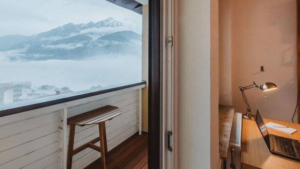 A room with a view of mountains through a window, a desk with a laptop, and a chair.
