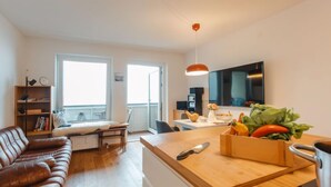 A modern living room with a brown leather sofa, a wooden dining table with a fruit bowl, and a kitchen area with a TV and a red pendant light.