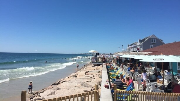 Ein Restaurant direkt am Strand mit Außenbereich, Blick auf das Meer und einem Gebäude im Hintergrund.