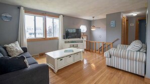 Ein Wohnzimmer mit einer Couch, einer gestreiften Couch, einem weißen Couchtisch, einem Fernseher auf einem Schrank und einer Treppe, die in einen anderen Raum führt.