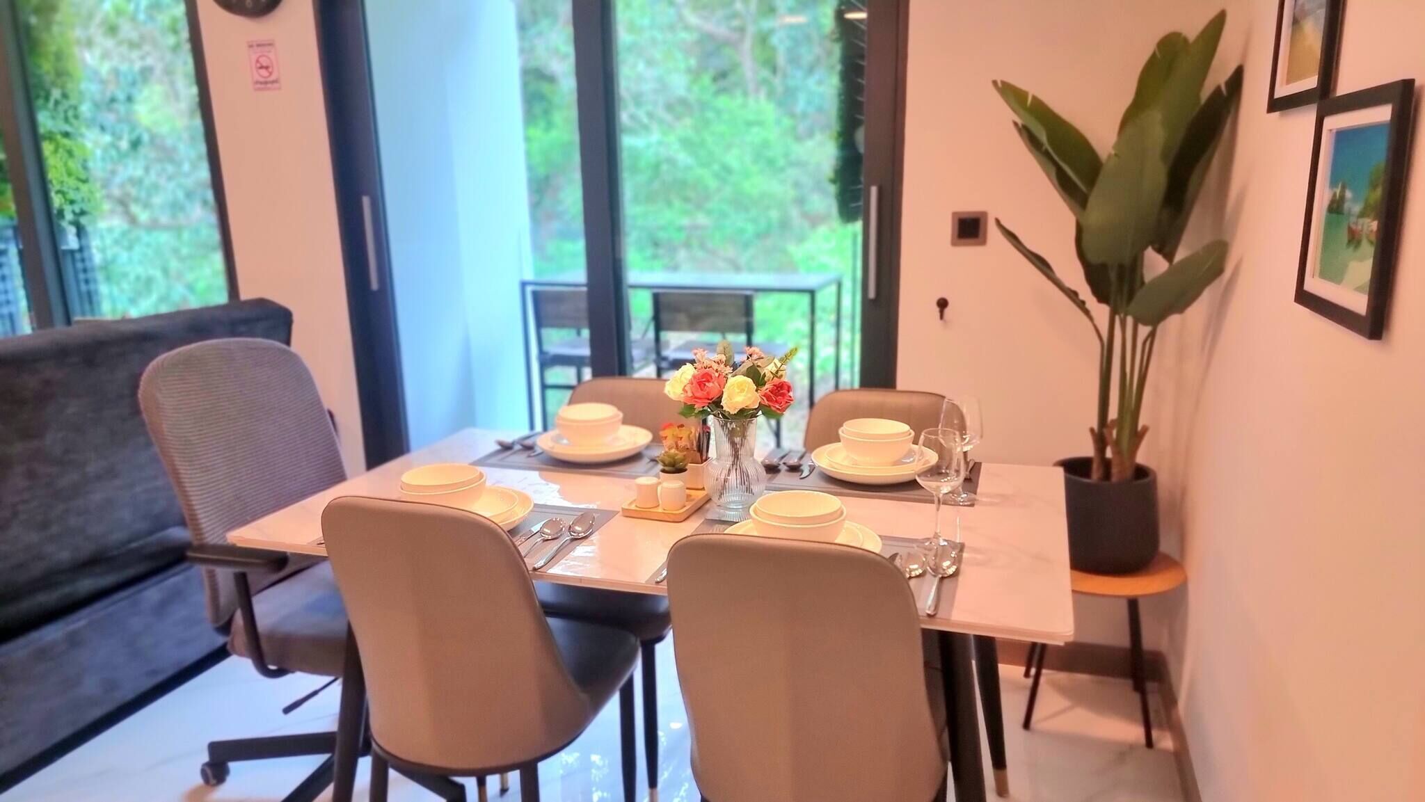 Uma sala de jantar com mesa posta para quatro, cadeiras, um vaso de flores e uma planta em vaso.