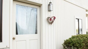 Une porte blanche avec une fenêtre, un paillasson et un petit jardin agrémenté d’un arbuste.