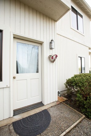A white door with a window, a doormat, and a small garden area with a bush.