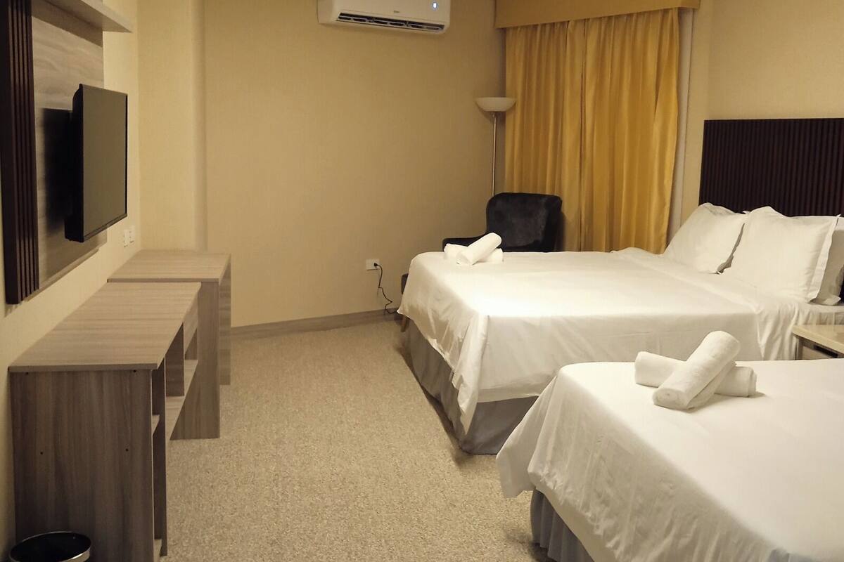 A hotel room with two beds, a desk, a chair, an air conditioning unit, and a TV.