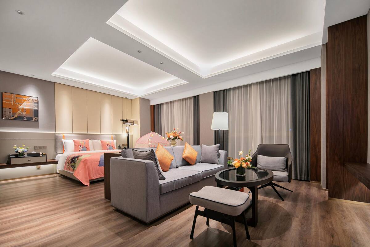 A modern hotel room with a sofa, a small table, a chair, a bed, and a large window with curtains.