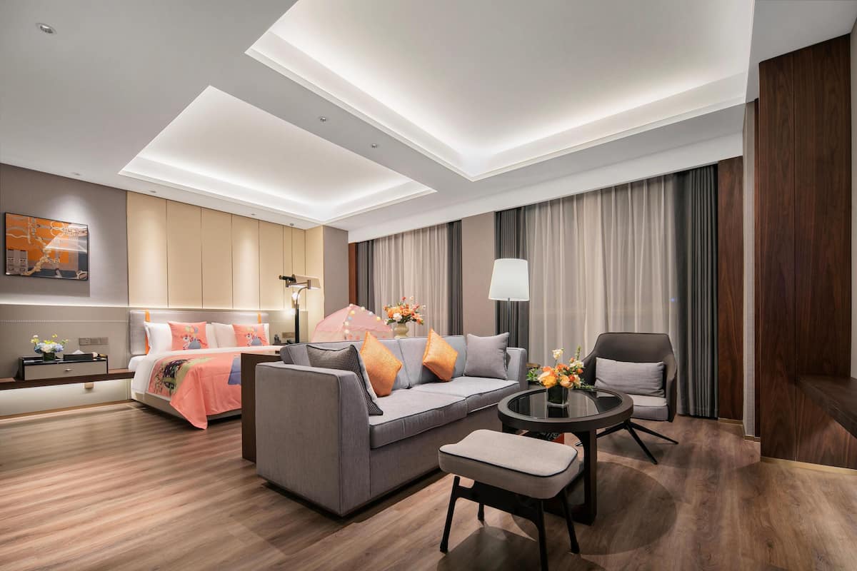 Quarto de hotel moderno com sofá, uma mesinha, cadeira, cama e uma janela grande com cortinas.