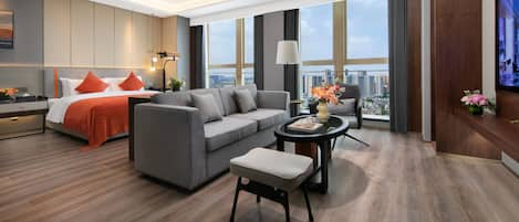 A modern hotel room with a bed, sofa, armchair, and a small table with a vase of flowers.