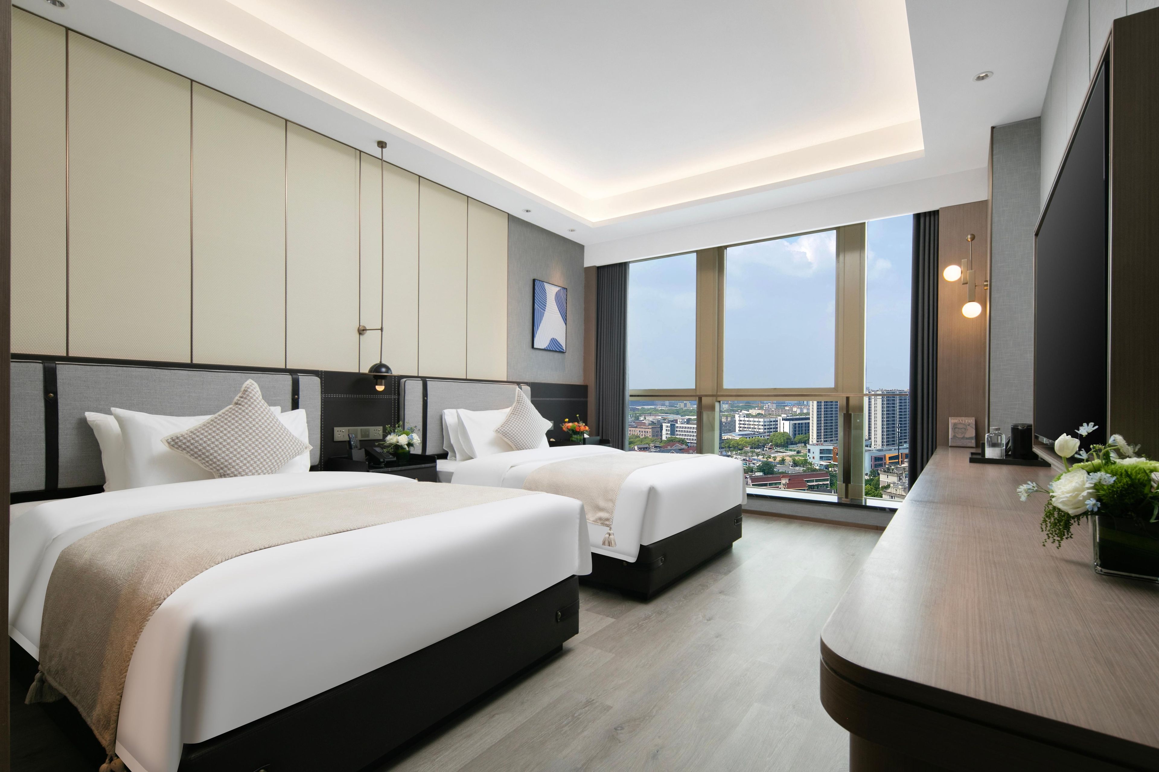 A modern hotel room with two beds, a large window with a city view, and a wooden desk with a vase of flowers.