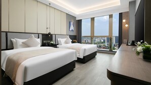 Een moderne hotelkamer met twee bedden, een groot raam met uitzicht op de stad en een houten bureau met een bloevas.