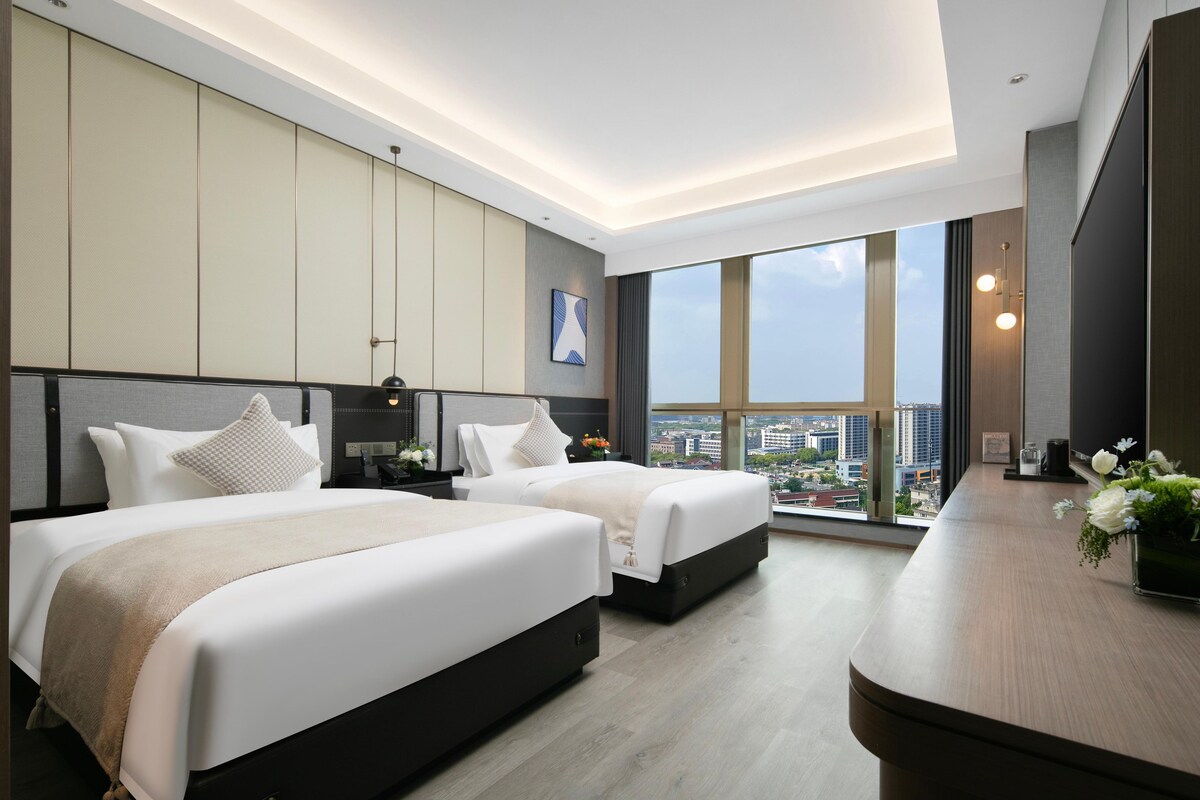 Una habitación de hotel moderna con dos camas, un ventanal grande con vistas a la ciudad y un escritorio de madera con un florero.