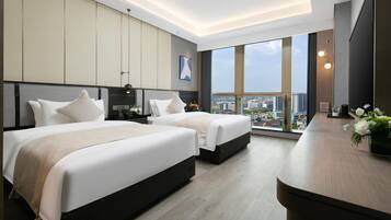 A modern hotel room with two beds, a large window with a city view, and a wooden desk with a vase of flowers.