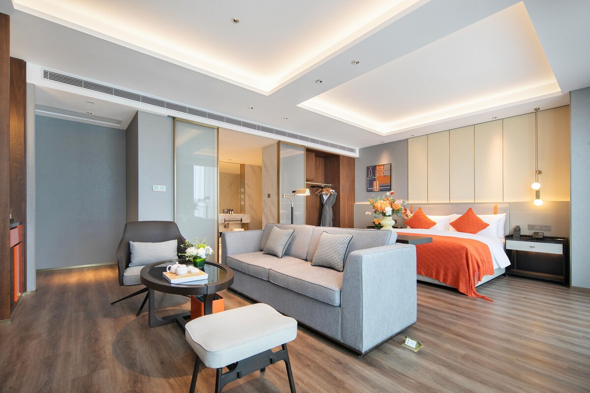Ein modernes Hotelzimmer mit einer Couch, einem Sessel, einem kleinen Tisch, einem Bett mit orangefarbenen Kissen und einem Nachttisch.