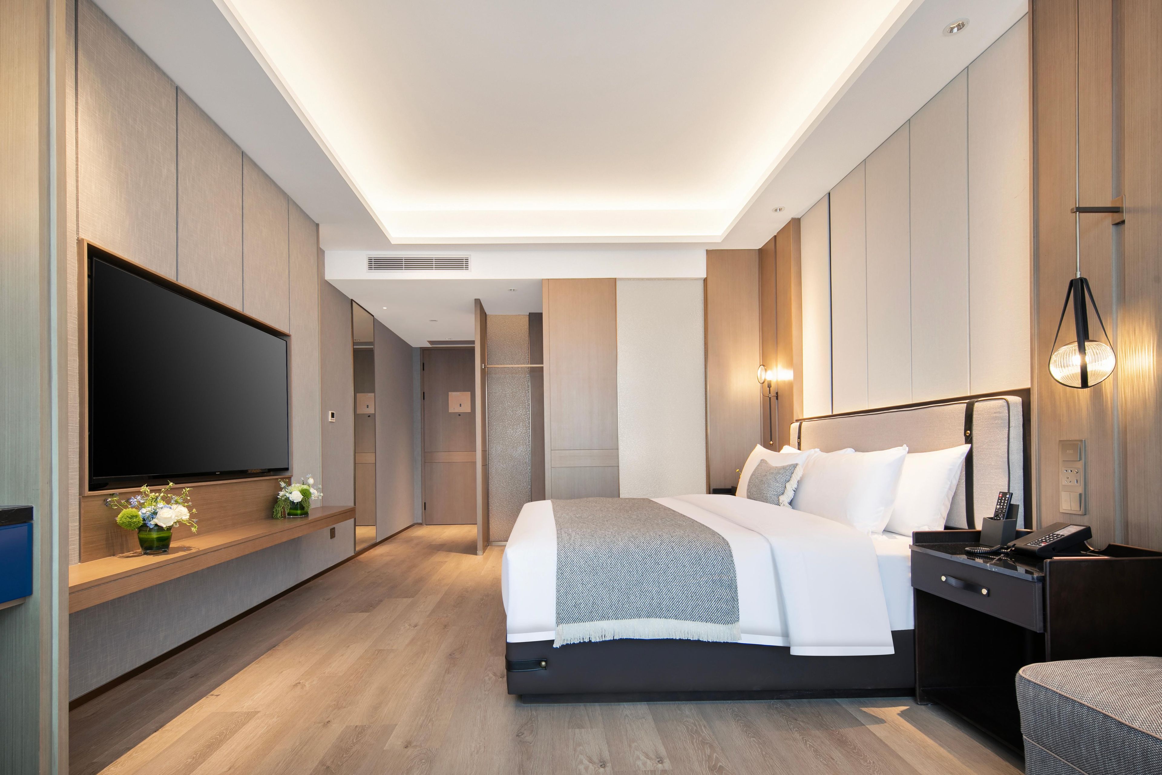 A modern hotel room with a large flat-screen TV, a bed with white linens, a bedside table with a lamp, and a wooden floor.