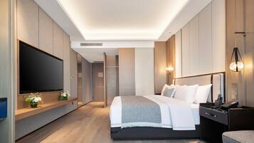 A modern hotel room with a large flat-screen TV, a bed with white linens, a bedside table with a lamp, and a wooden floor.